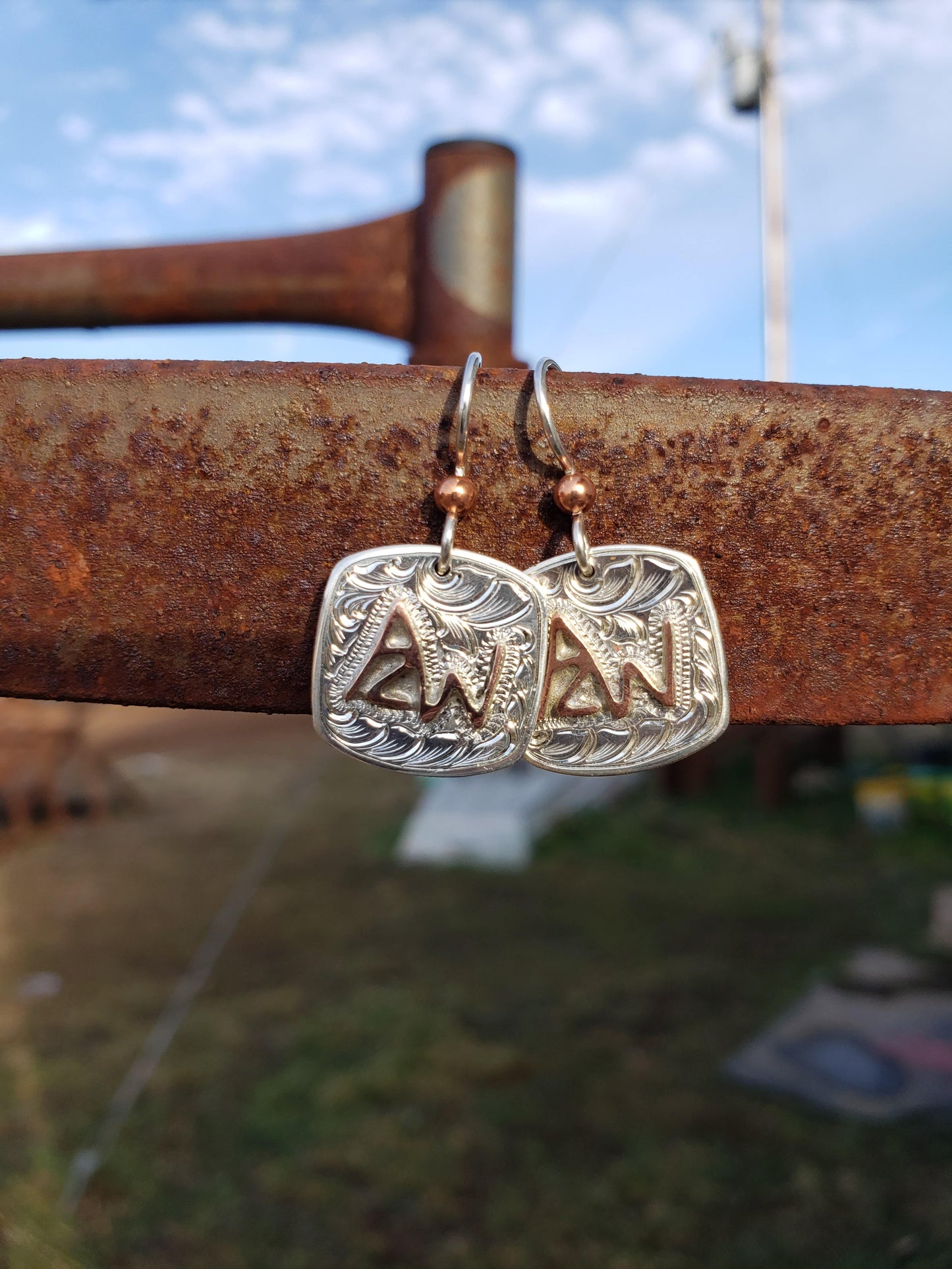A pair of hand-made, engraved sterling silver earrings featuring a cattle brand design, displayed against an outdoor backdrop.