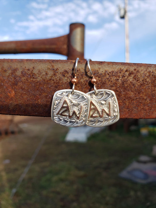 A pair of hand-made, engraved sterling silver earrings featuring a cattle brand design, displayed against an outdoor backdrop.